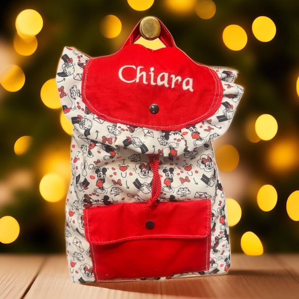 Zainetto bianco con Minnie Mouse e nome Chiara ricamato su sfondo di luci natalizie sfocate.